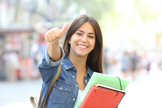 A happy student with booklets in her arm gives the thumbs up