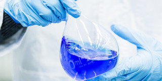 Two hands in blue rubber gloves hold a volumetric flask filled with blue liquid.