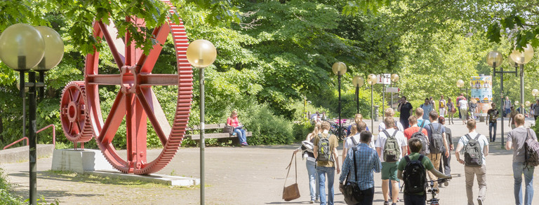 Header-Bild: Studenten, rote Zahnräder (Campus Nord) Studenten im Sommer auf dem Campus Nord, rote Zahnräder im Hintergrund