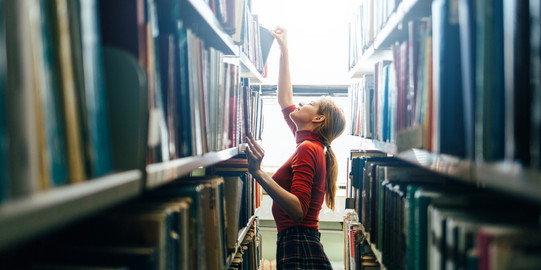 Junge Frau am Bücherregal Junge Frau sucht Bücher am Regal und zieht eins heraus