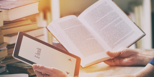 Mann sitzt mit E-Book-Reader und Buch am Tisch Ein Mann sitzt mit einem E-Book-Reader und einem Buch in den Händen an einem Tisch mit Büchern, Schreibzeug und Papieren