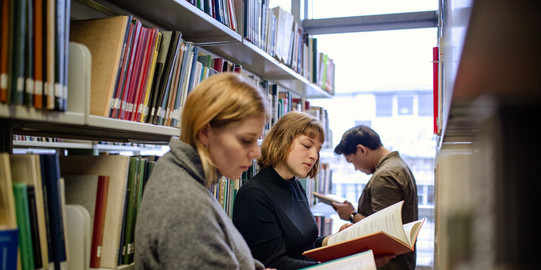 Students stand reading in front of bookshelves in the library.
