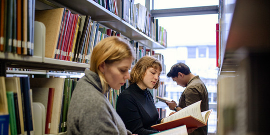 Bücherrecherche in der Bibliothek Studierende stehen lesend vor Bücherregalen in der Bibliothek.